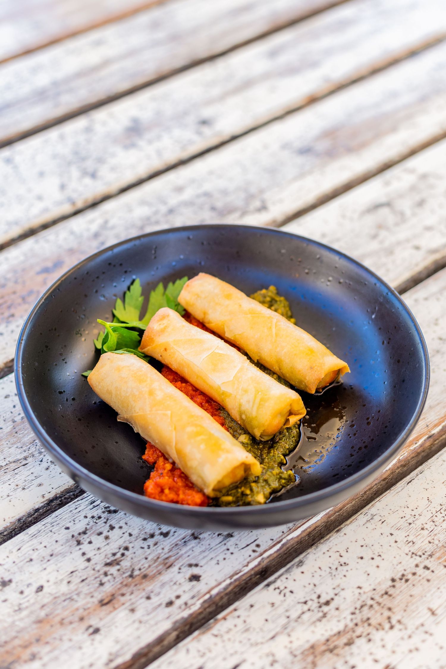 Golden crispy spring rolls served with sauce and garnish on a black plate.