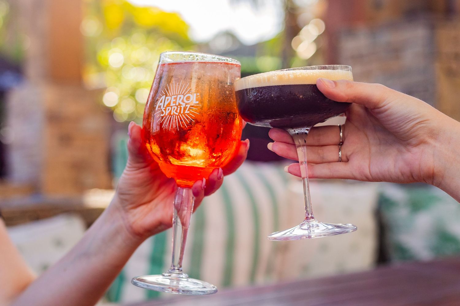 Two hands toasting with colorful cocktails against a blurred outdoor background.