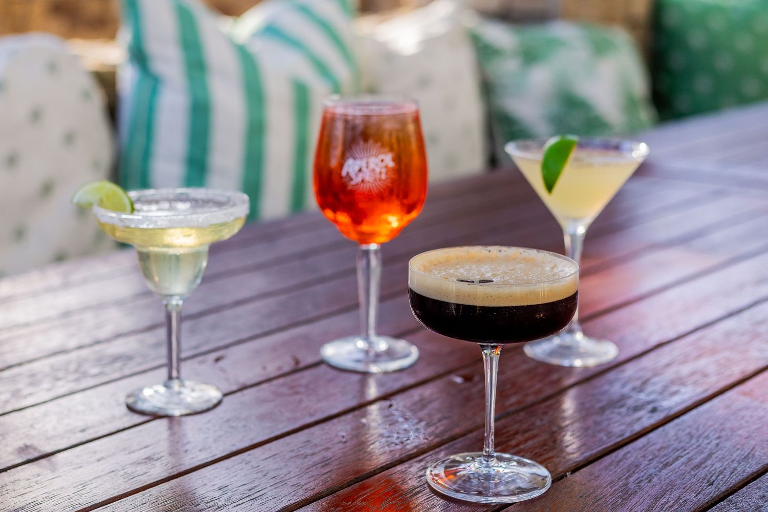 Three elegant cocktails including a margarita, aperol spritz, and espresso martini on wooden table.