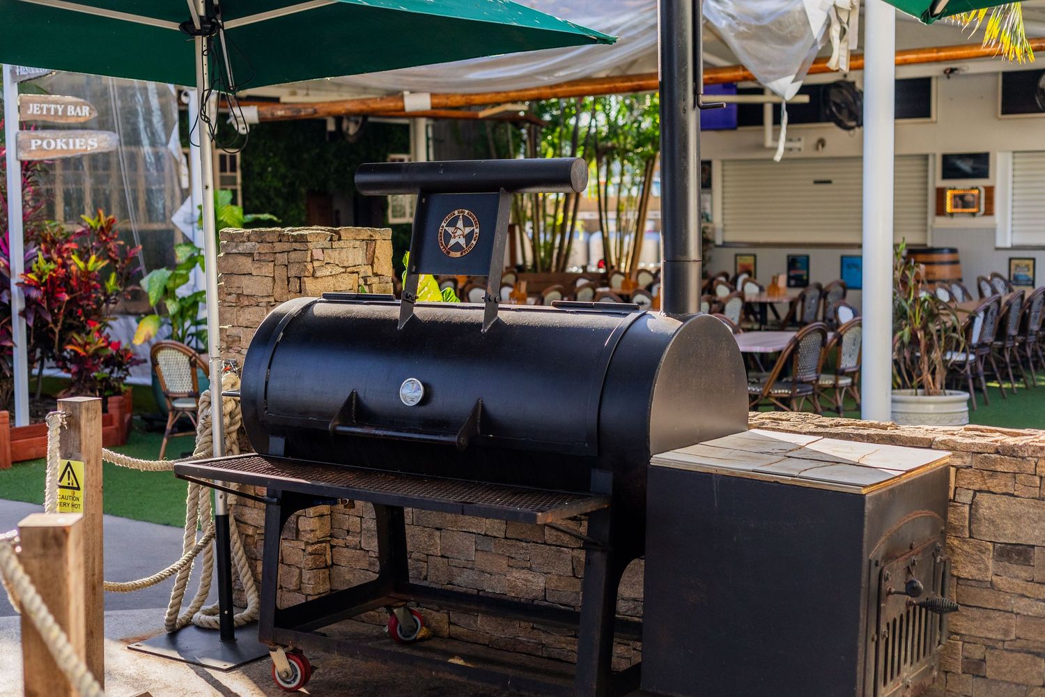 Large black smoker grill on stone patio under green market umbrella.