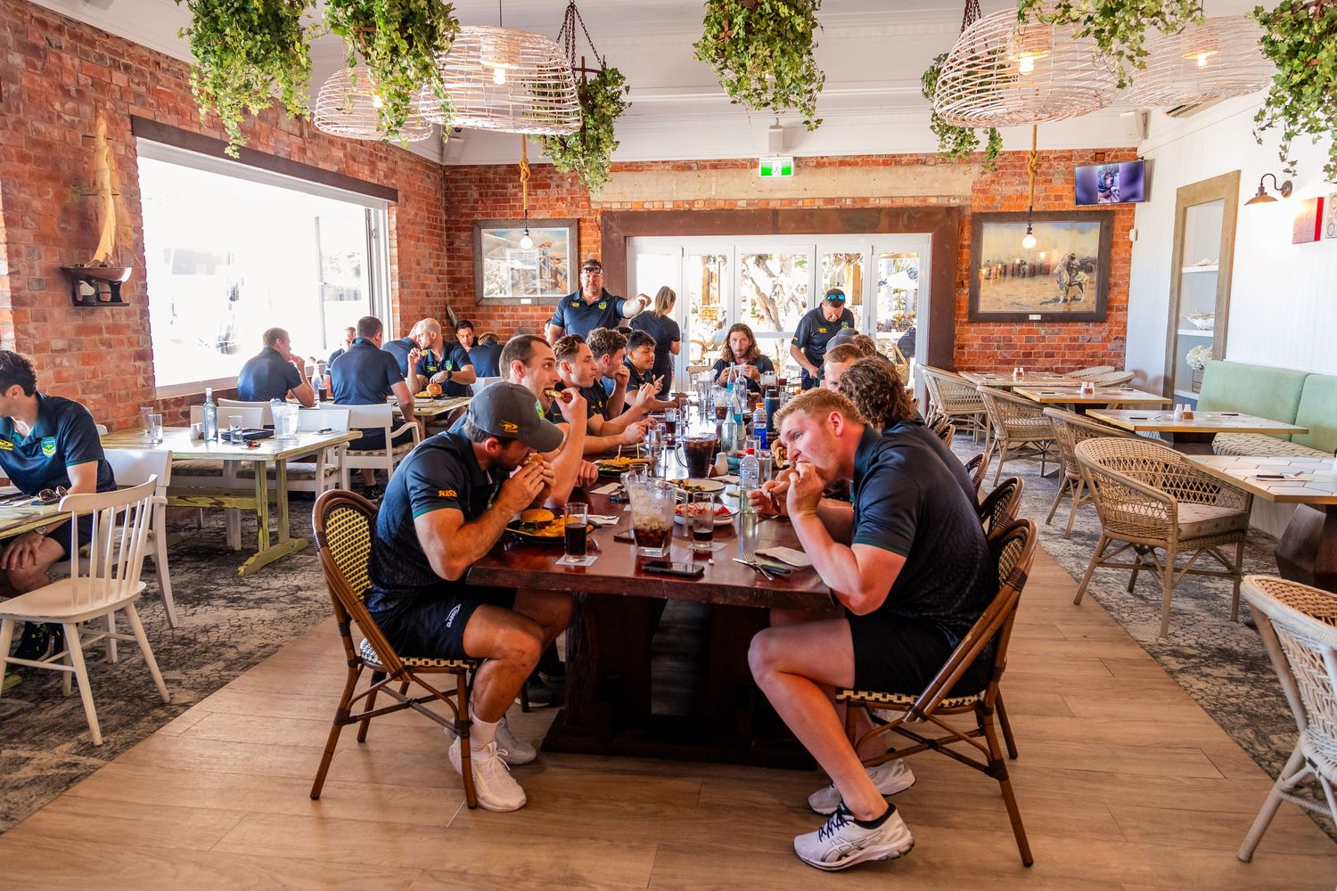 Diners enjoying their meals in a bright rustic restaurant with exposed brick walls and hanging plants.
