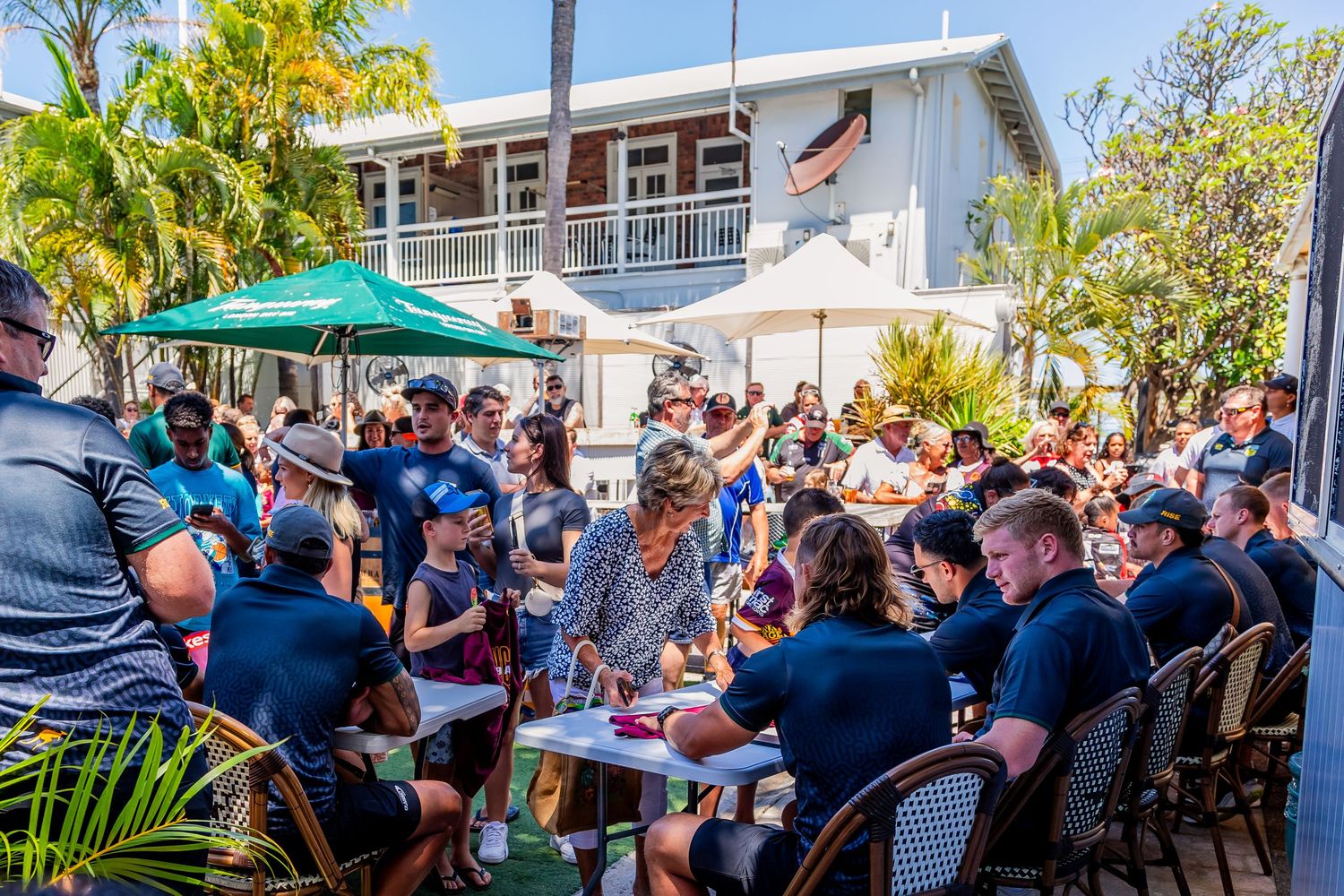 Large crowd gathered at an outdoor patio dining area under turquoise umbrellas and palm trees.
