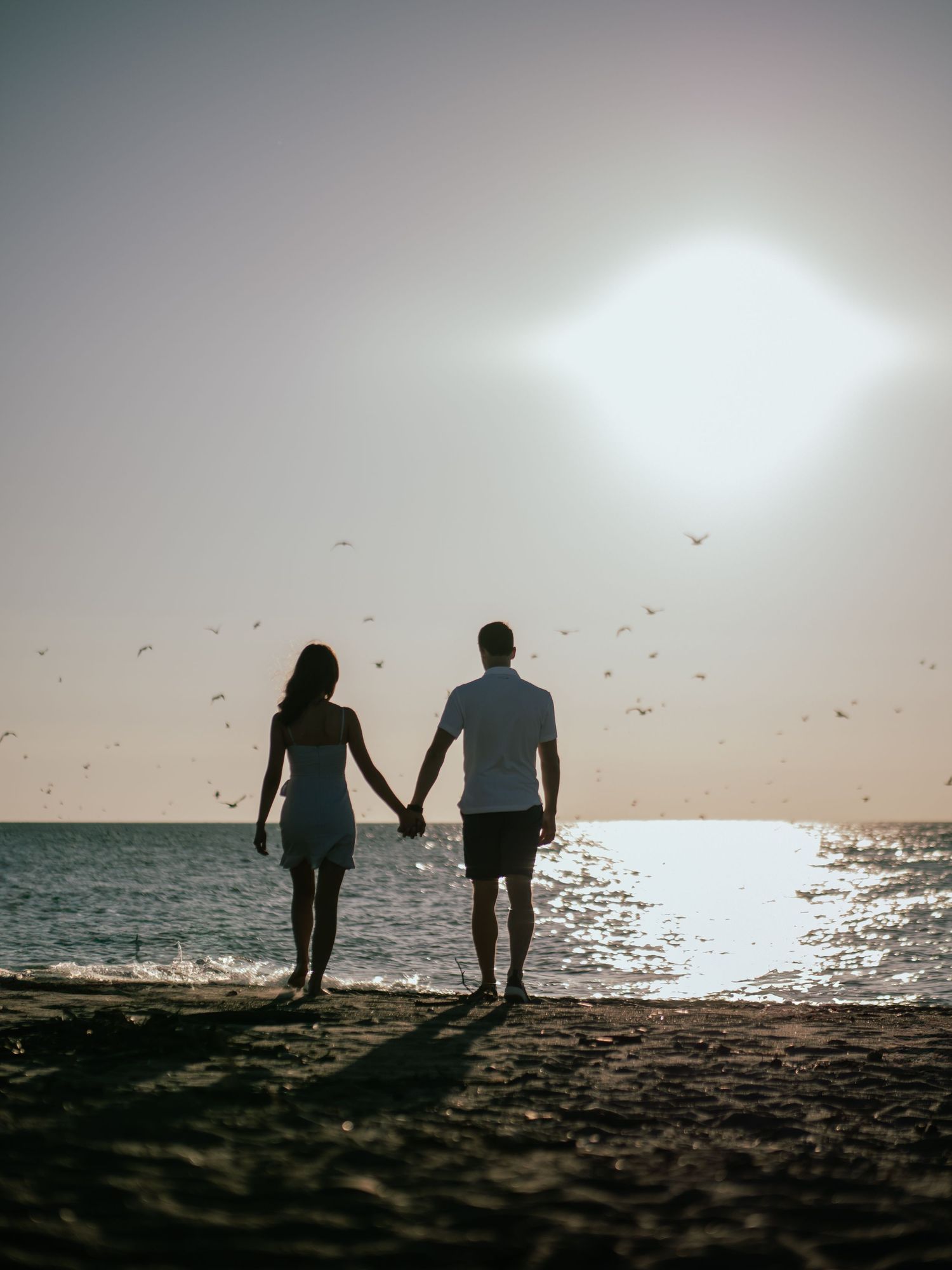 Silhouetted couple holding hands on beach at sunset with birds flying overhead.