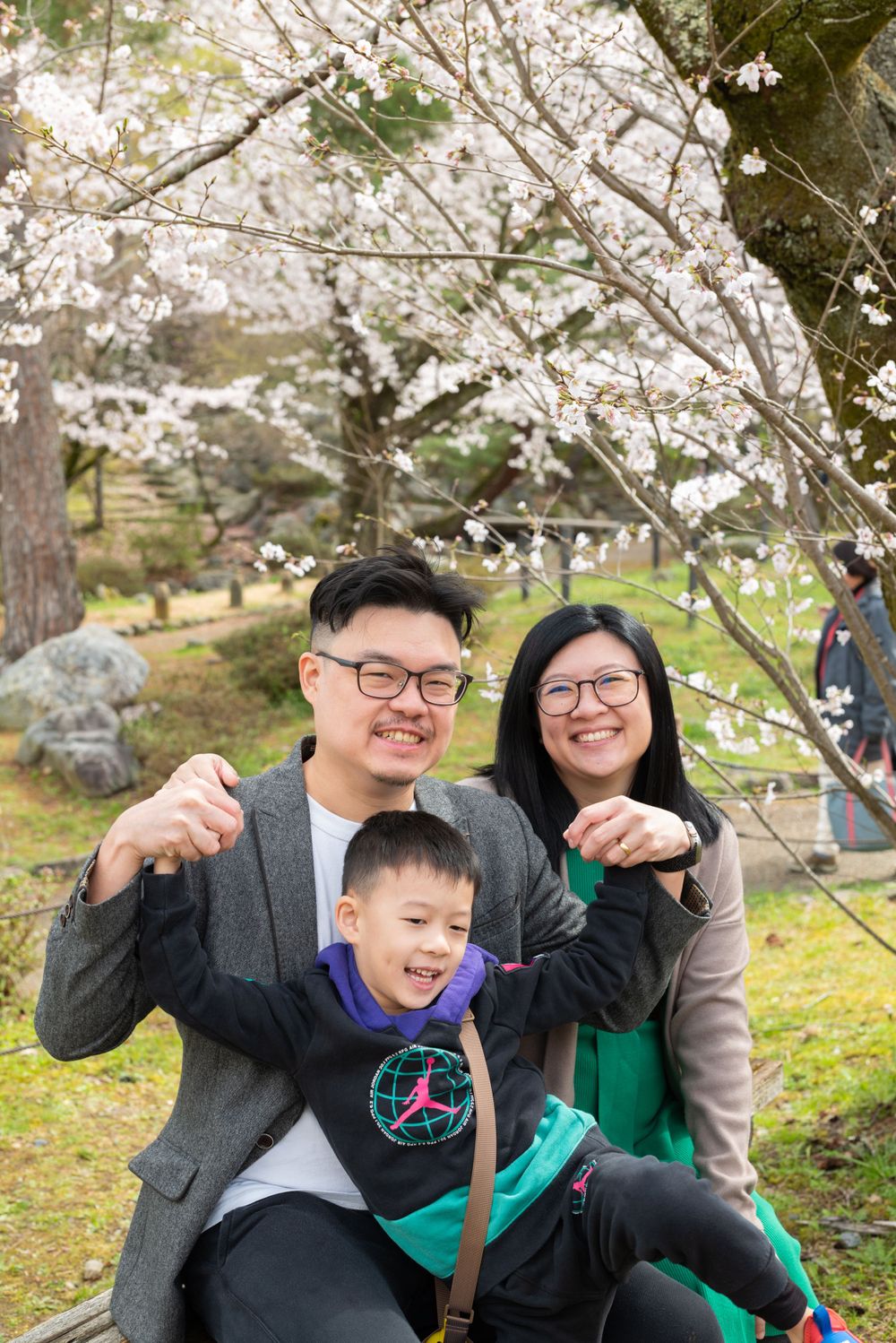 Smiling Asian family posing playfully under white cherry blossoms in a scenic park.