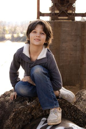 Young child crouches on rocks near industrial equipment during a sunny outdoor photoshoot.