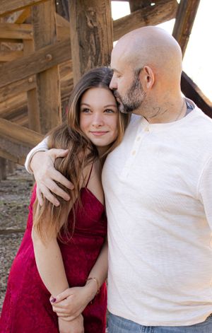 Couple embracing outdoors, wearing white and red outfits against rustic wooden structure.