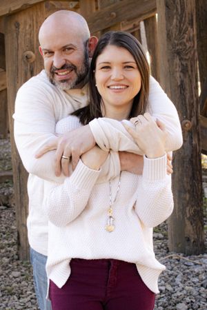 Couple in matching white sweaters share a warm embrace against wooden backdrop.