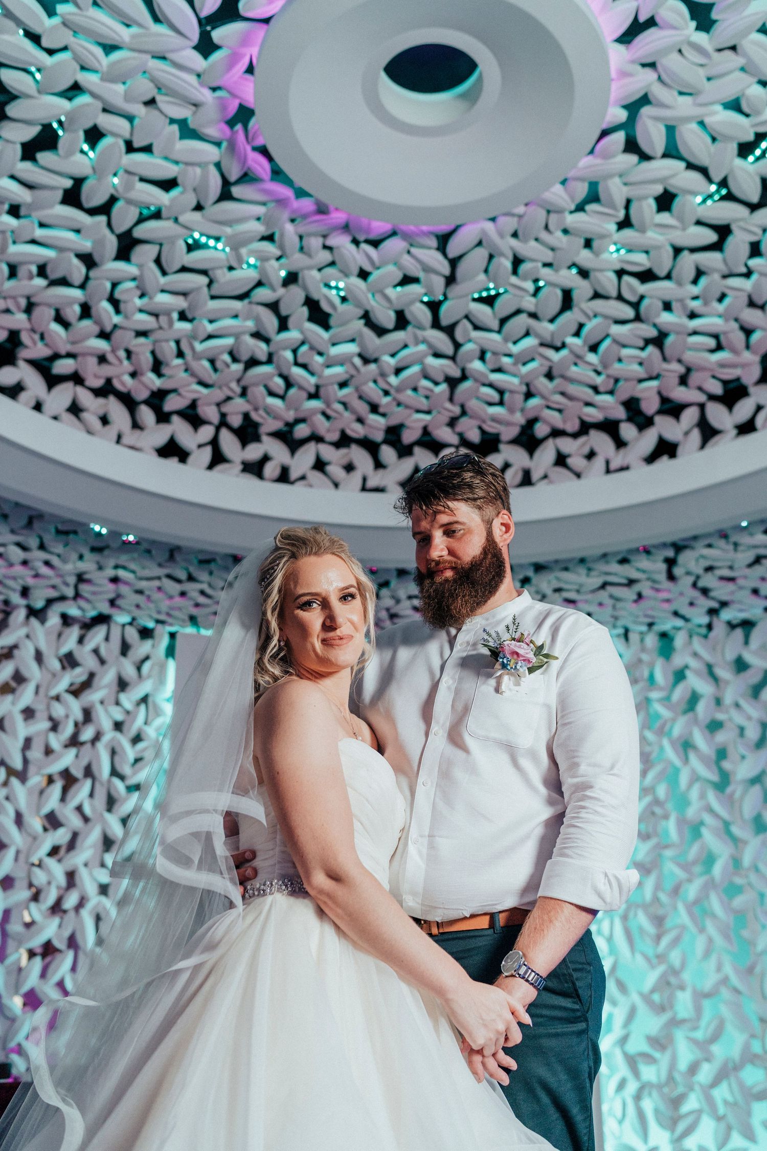Newlyweds pose under an ornate ceiling with intricate geometric patterns and blue-tinted lighting at their wedding reception.