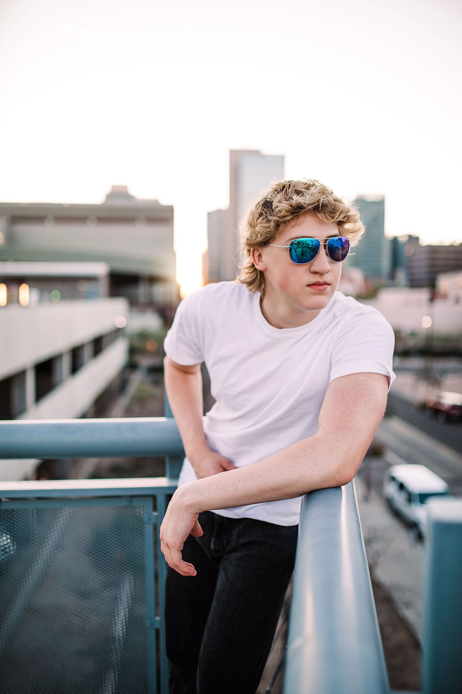 Stylish young man in white t-shirt and blue mirrored sunglasses leaning on urban rooftop railing.