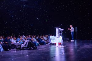 Speaker presenting to a seated audience in a conference hall with starry lighting effects.