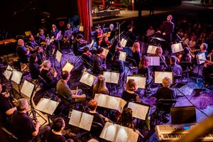 Full orchestra performing on stage with illuminated music stands and red curtain backdrop.