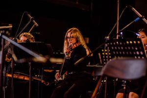 Orchestra musician performing on stage with music stands and equipment visible in dark lighting.