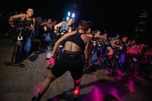 Group fitness class with people on spin bikes in dark room with neon lights.