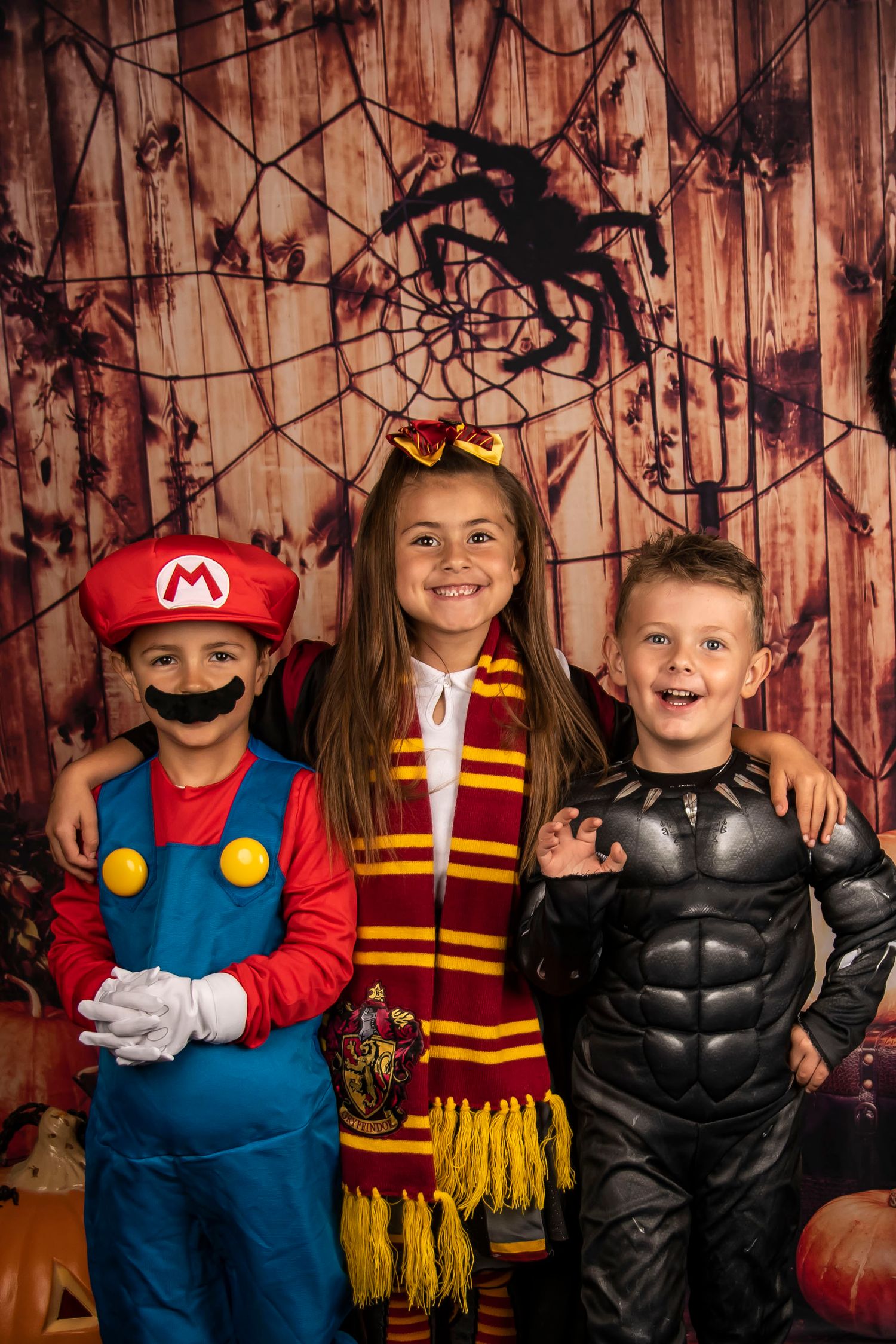 Three children in Mario, Harry Potter, and superhero costumes pose against spooky wooden backdrop.