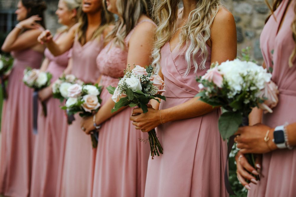 Bridesmaids in dusty rose gowns hold white floral bouquets while standing in a row at an outdoor wedding ceremony.