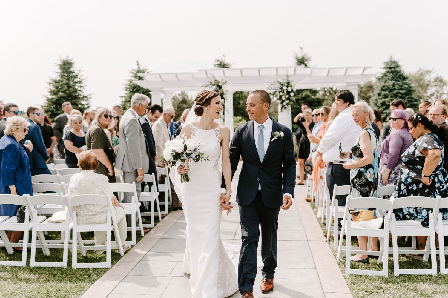 A wedding ceremony on a sunny day with guests seated in white chairs and a floral arch backdrop.