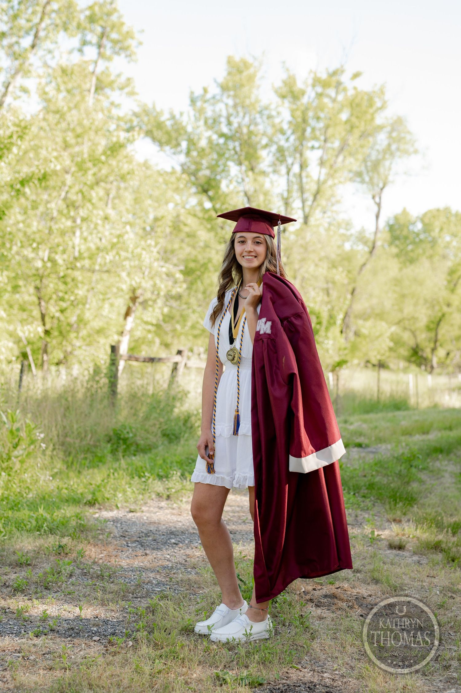 Graduate in maroon graduation regalia and white dress poses on nature path with trees in background.