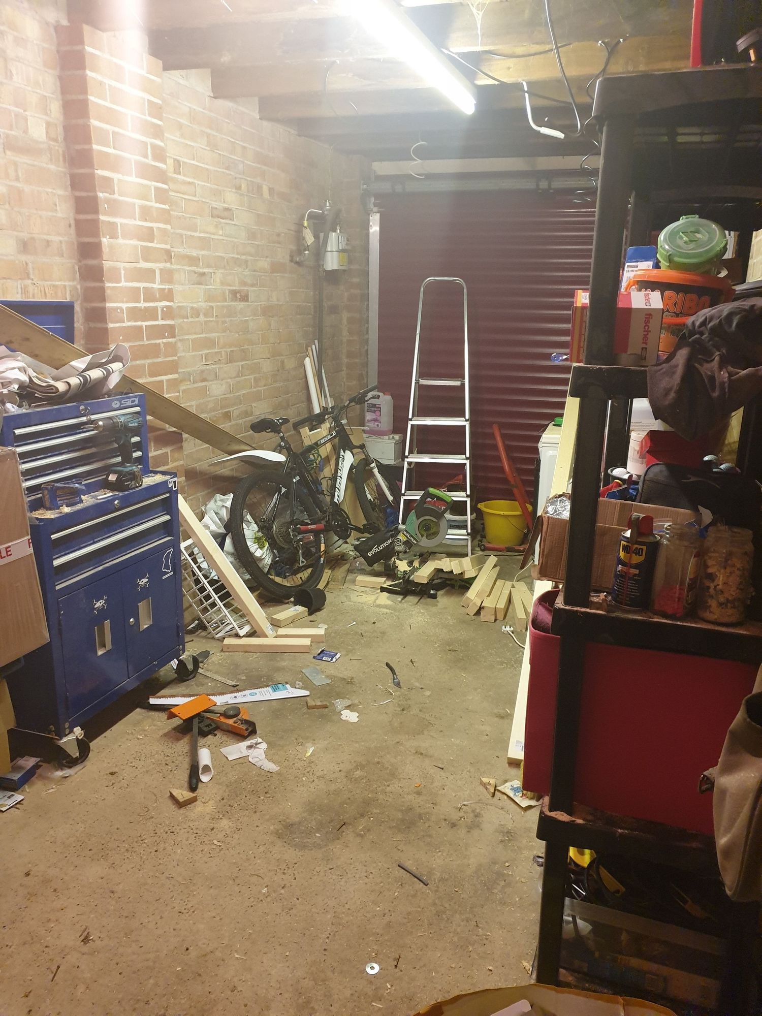 Messy garage workshop with tool chest, ladder, and various equipment scattered around brick walls.