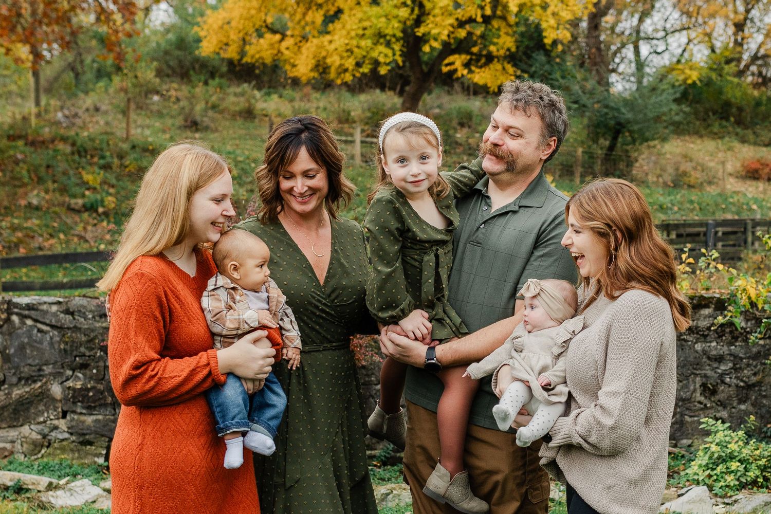Family gathering outdoors during autumn dressed in cozy sweaters with toddlers and infants against fall foliage.