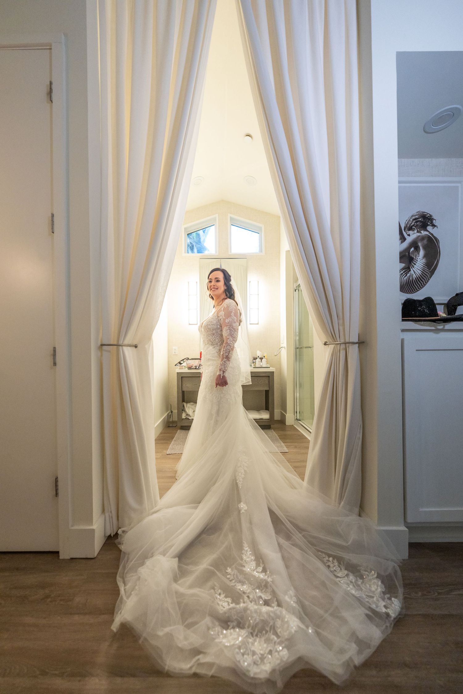 Wedding dress with long lace train displayed against white curtains in natural lighting.