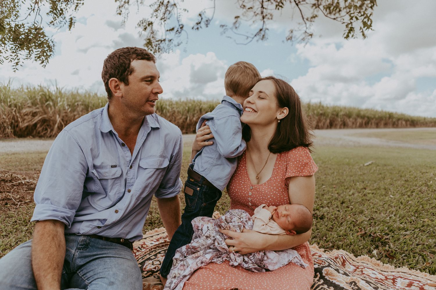 A family sits on a picnic blanket outdoors, parents smiling at their toddler while mother holds a newborn.