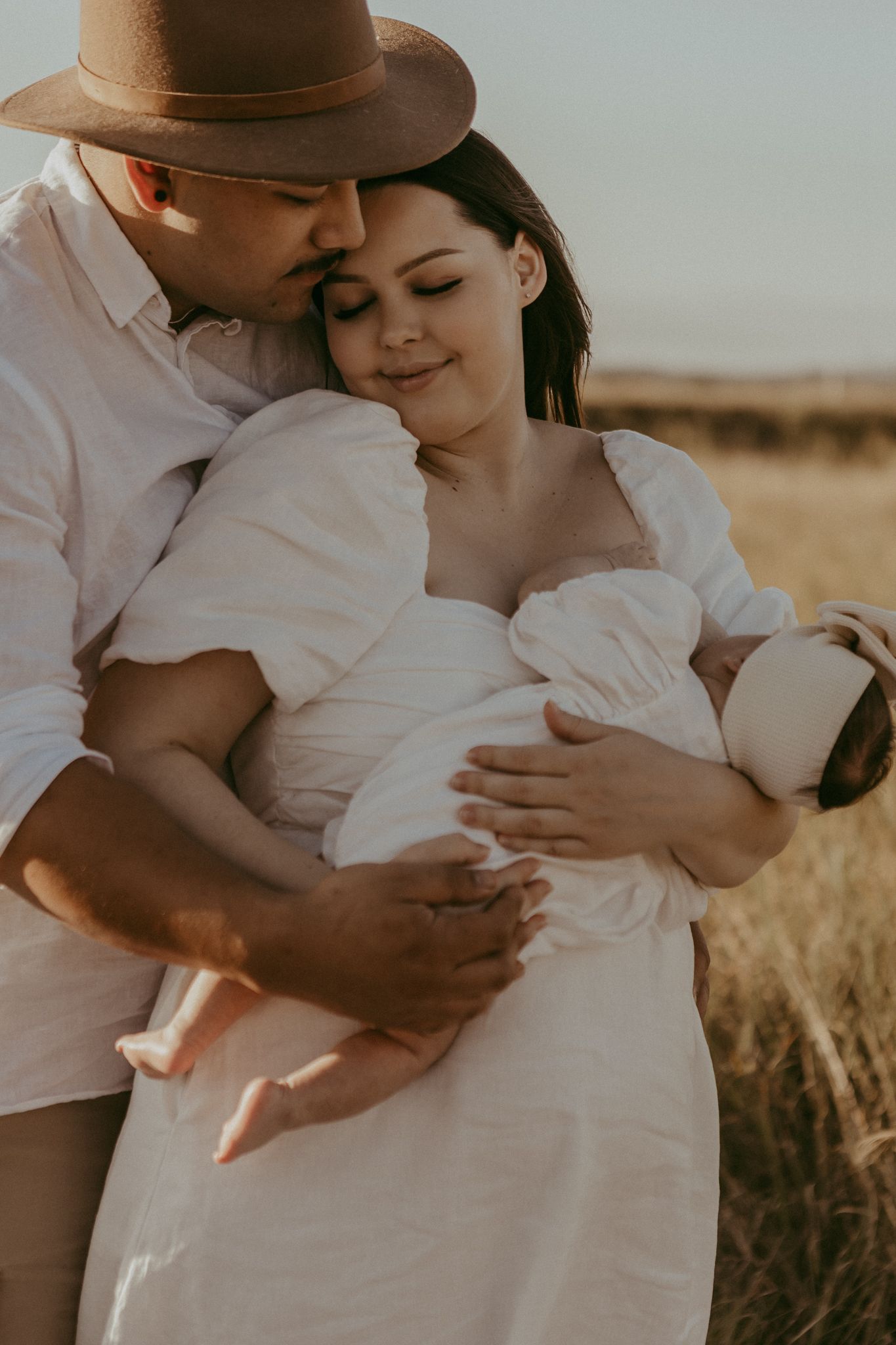 Couple embracing in golden field, pregnant woman in white dress with partner in brown hat at sunset.