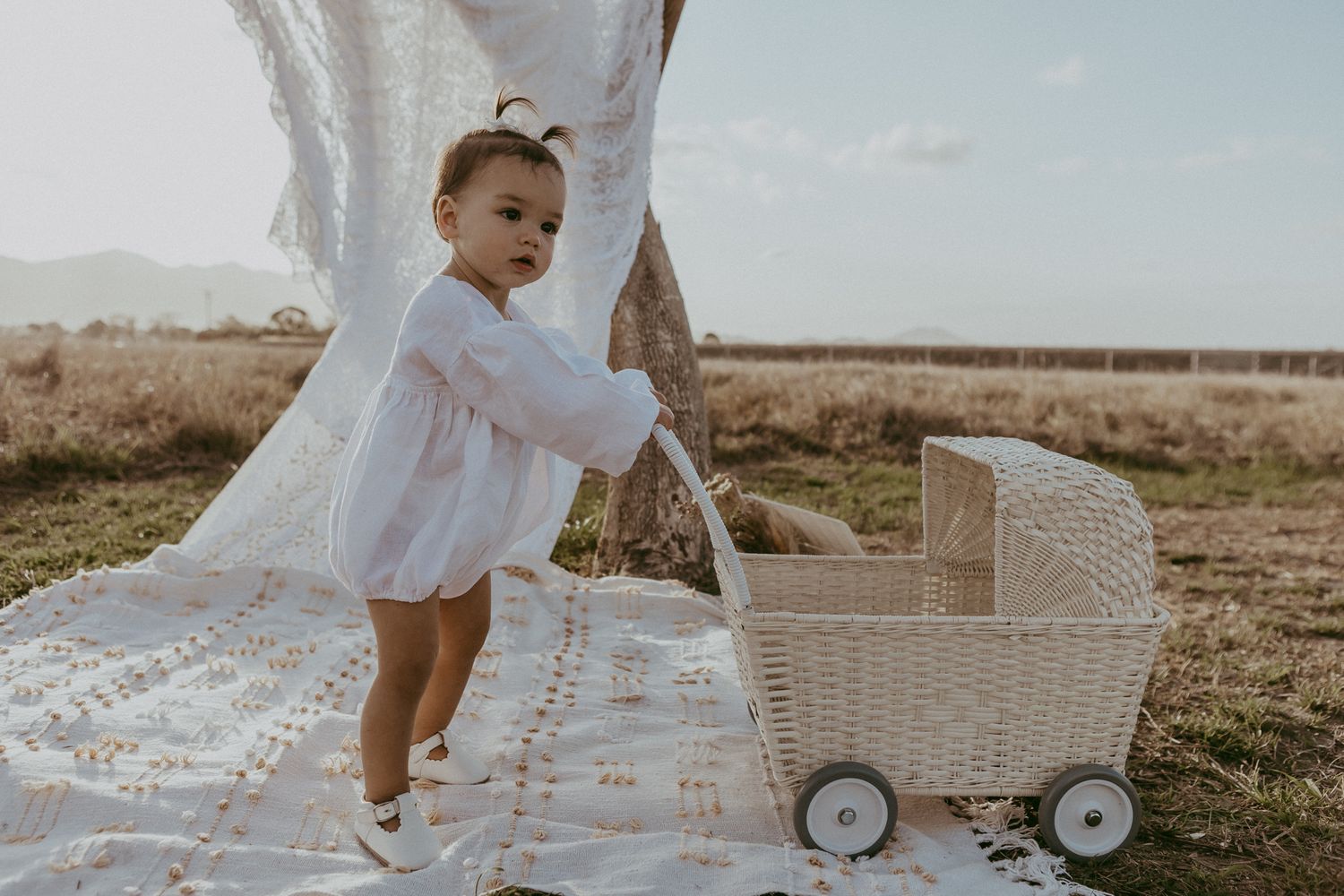 Baby in white romper pushes a wicker pram on a patterned blanket in a sunny field.