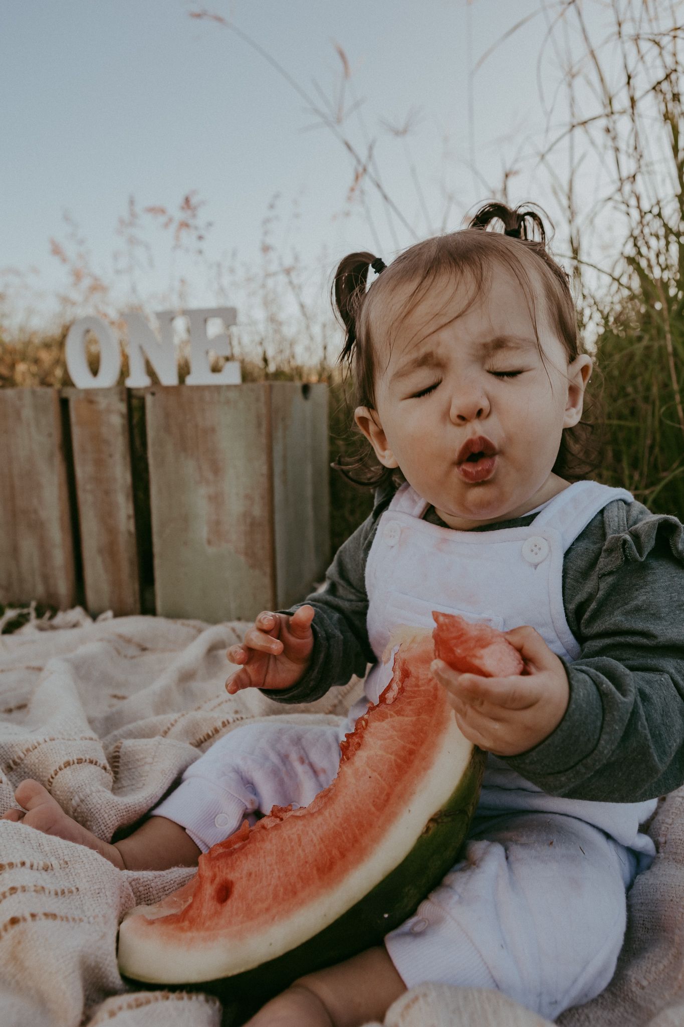 Adorable one-year-old baby girl eating watermelon slice outdoors at golden hour birthday session.