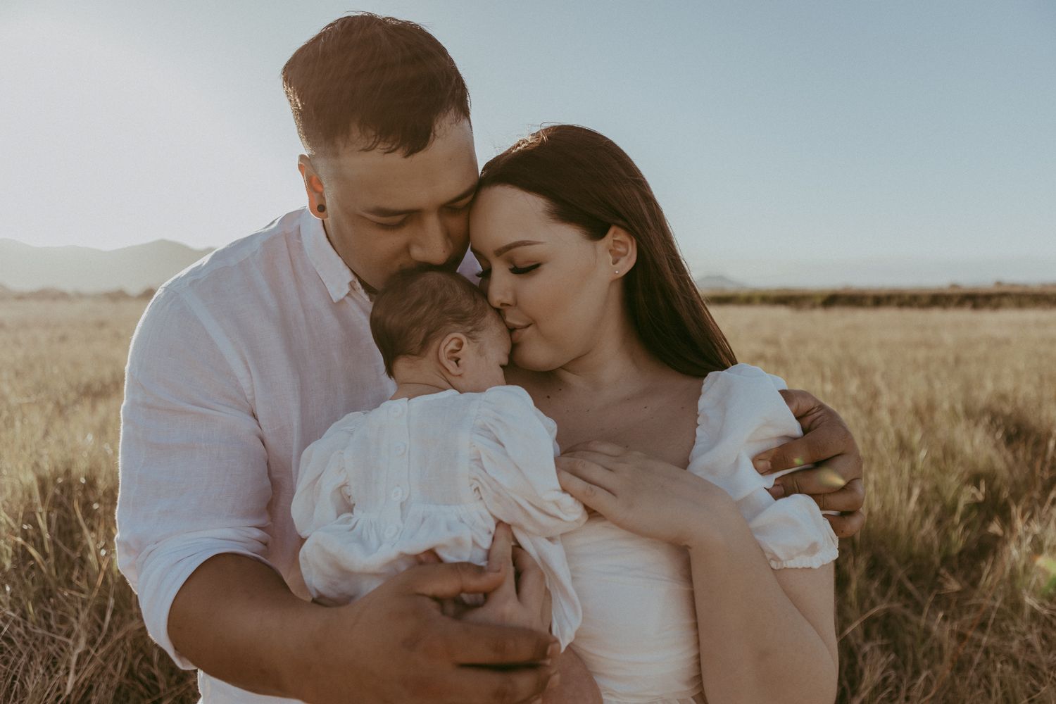 Parents tenderly holding their newborn baby in a golden sunlit wheat field at sunset.