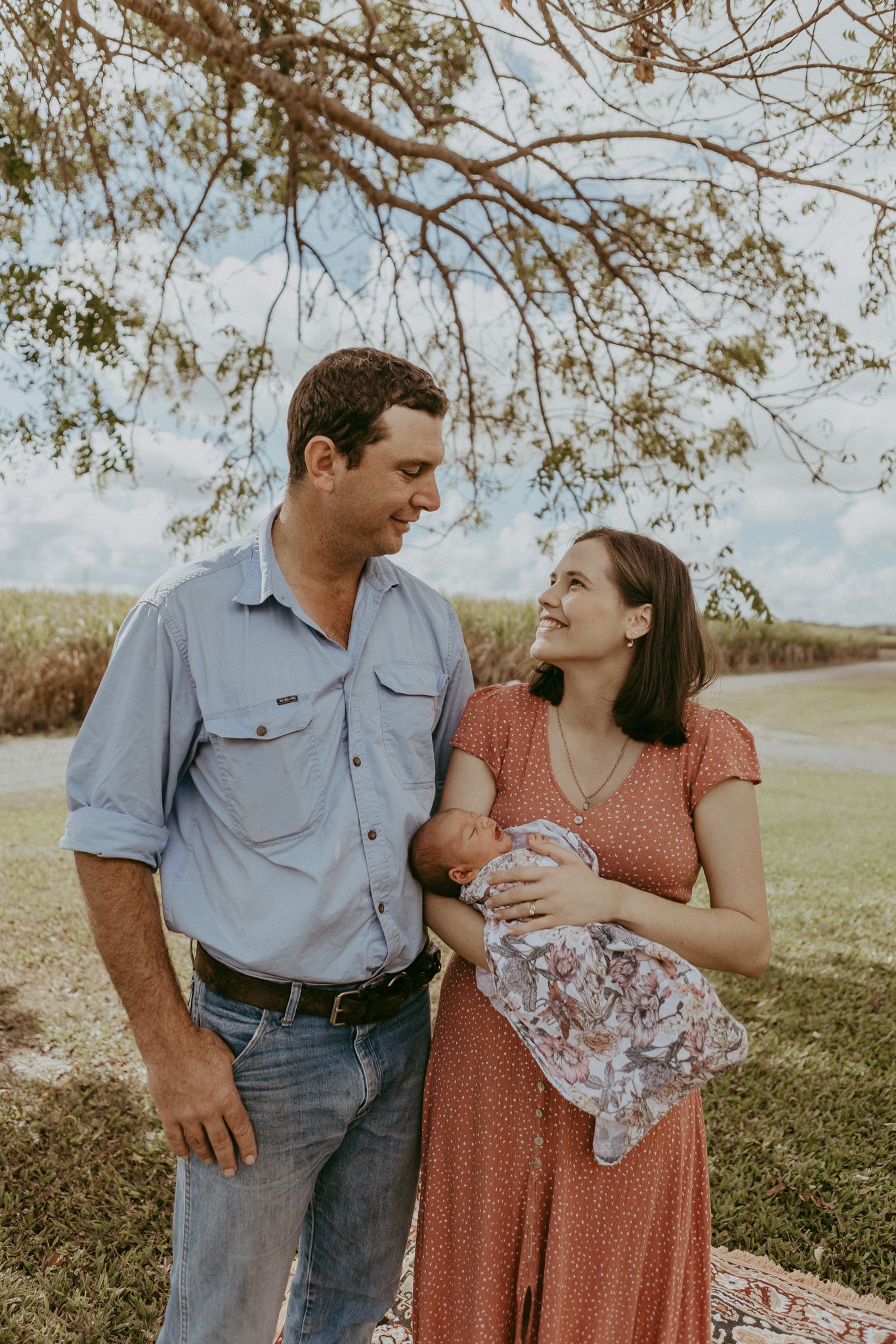 A couple stands outdoors under a tree, smiling at each other while the woman holds a newborn baby.