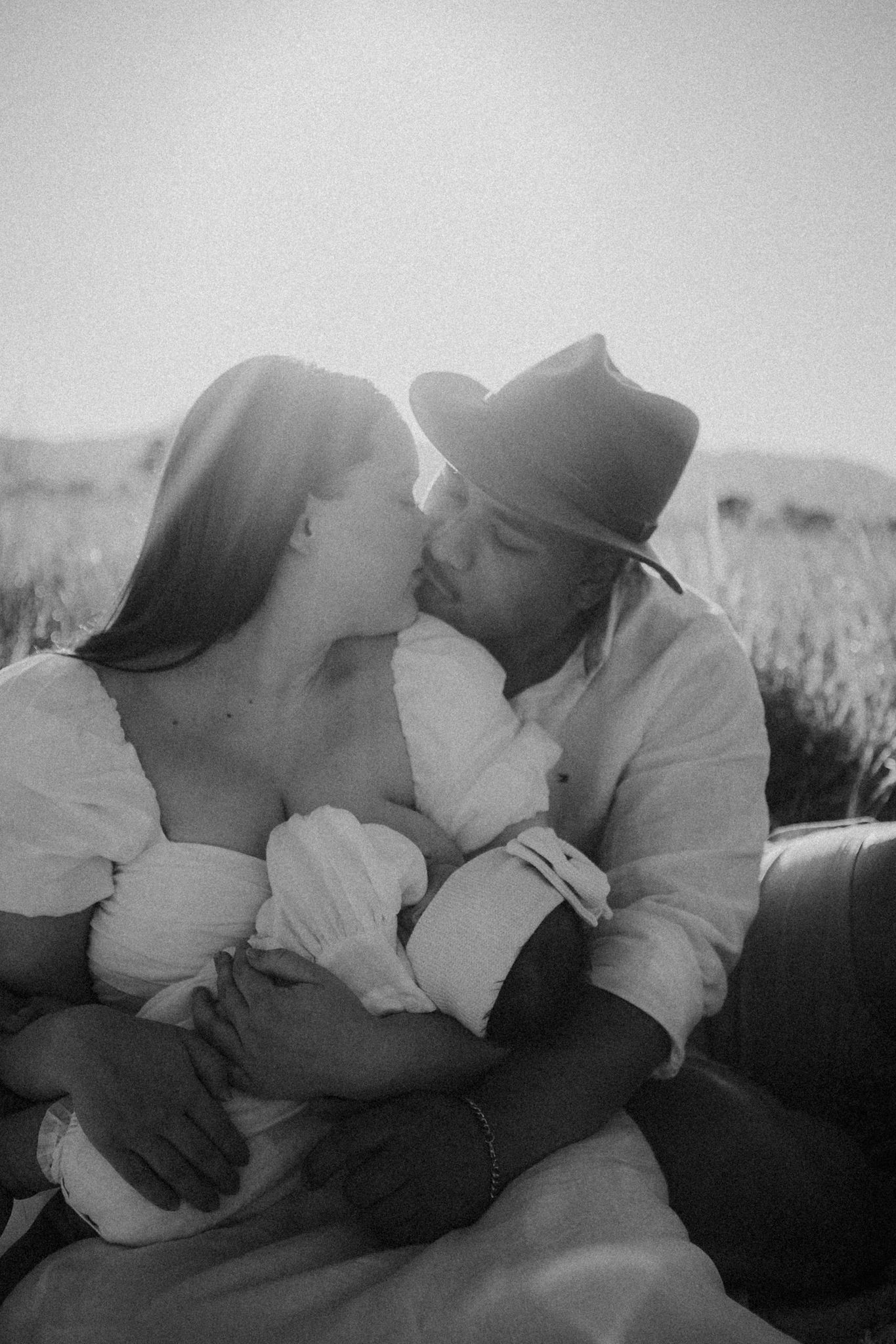 Black and white photo of a couple kissing while holding their newborn baby in a sunny field.