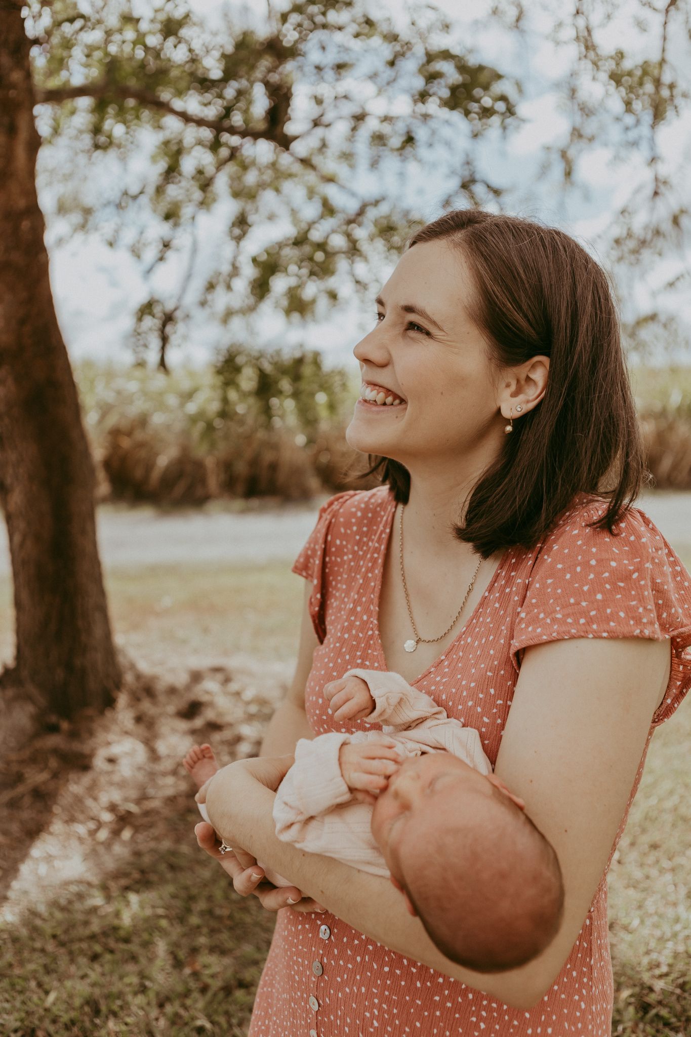 A smiling woman in a red polka dot dress holds her newborn baby outdoors beneath a tree.
