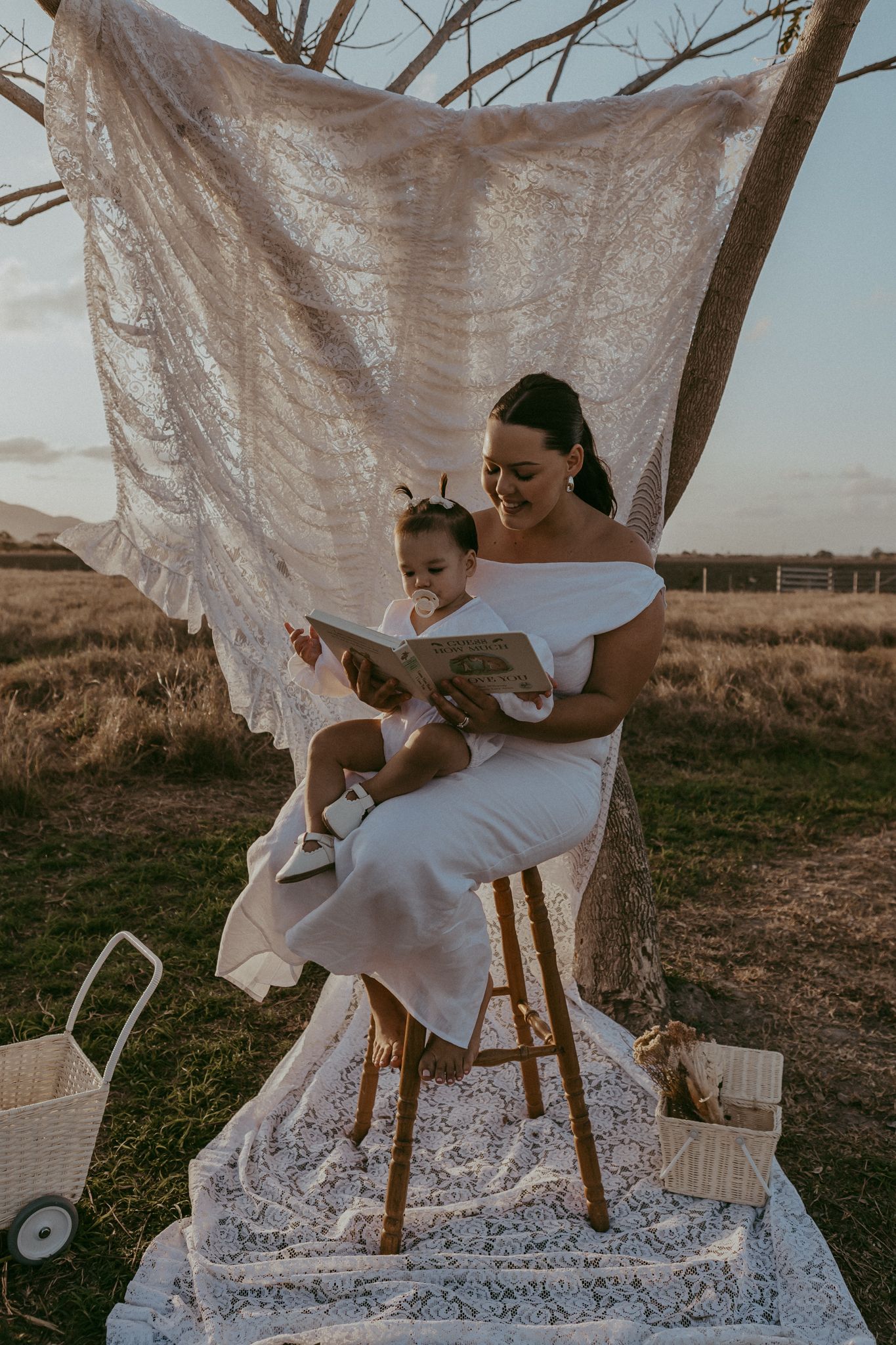 Mother reads a book to her toddler on a wooden stool in a golden field at sunset.