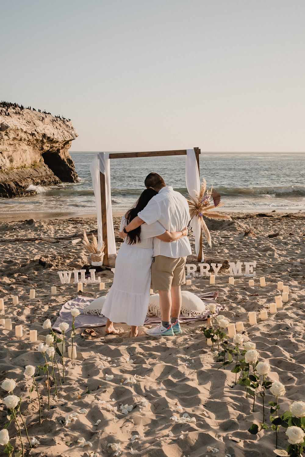 A couple embraces under a wooden arch during a sunset beach proposal.