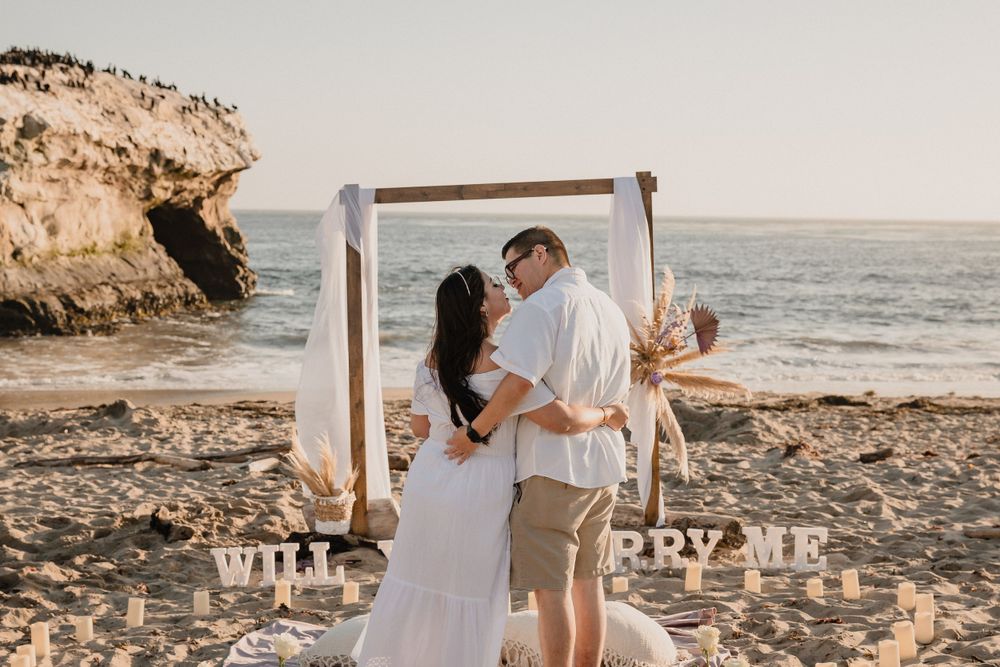 A joyful moment captured during a surprise beach proposal at sunset with decorative wooden arch.