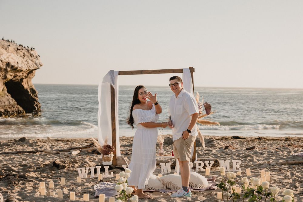 A romantic beach proposal setup with a wooden arch and candles at sunset.