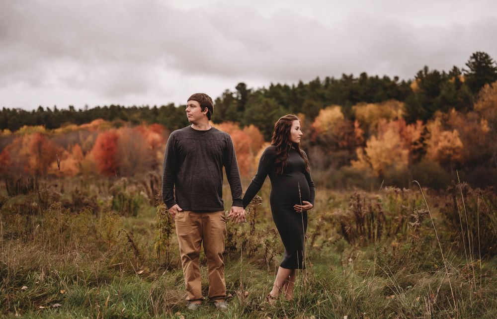 Couple holding hands while walking through autumn field with fall foliage in background.