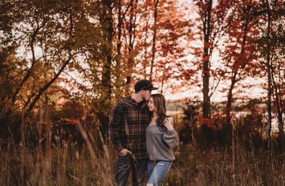 Romantic couple embraces in an autumn field during sunset surrounded by tall grass and trees.