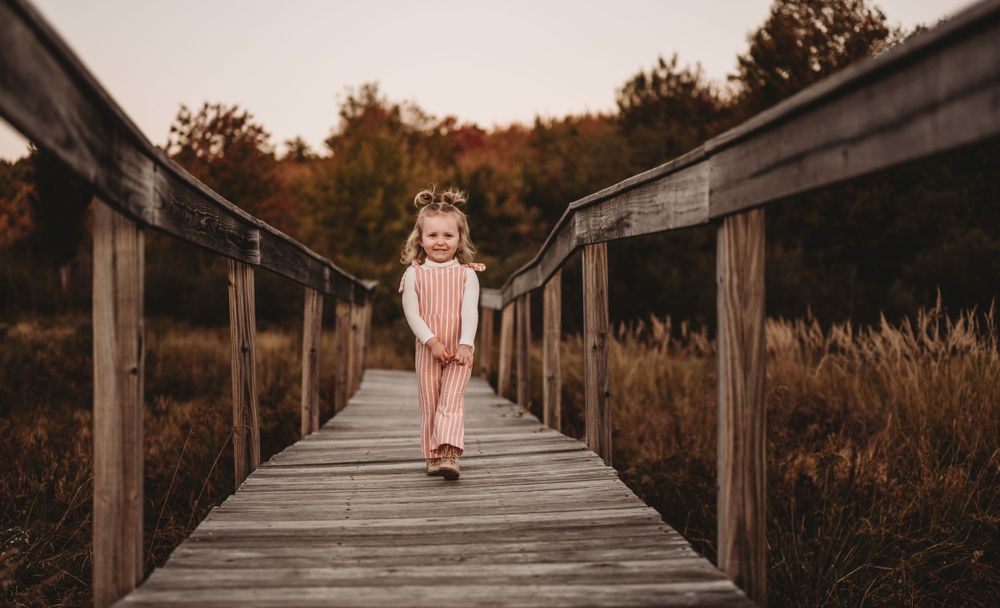 Child in pink dress walks along wooden boardwalk path through autumn marsh landscape.