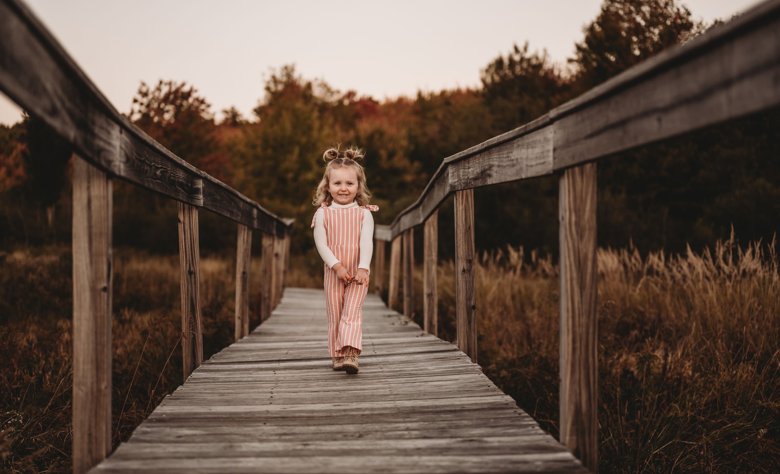 Child in pink dress walks along wooden boardwalk path through autumn marsh landscape.
