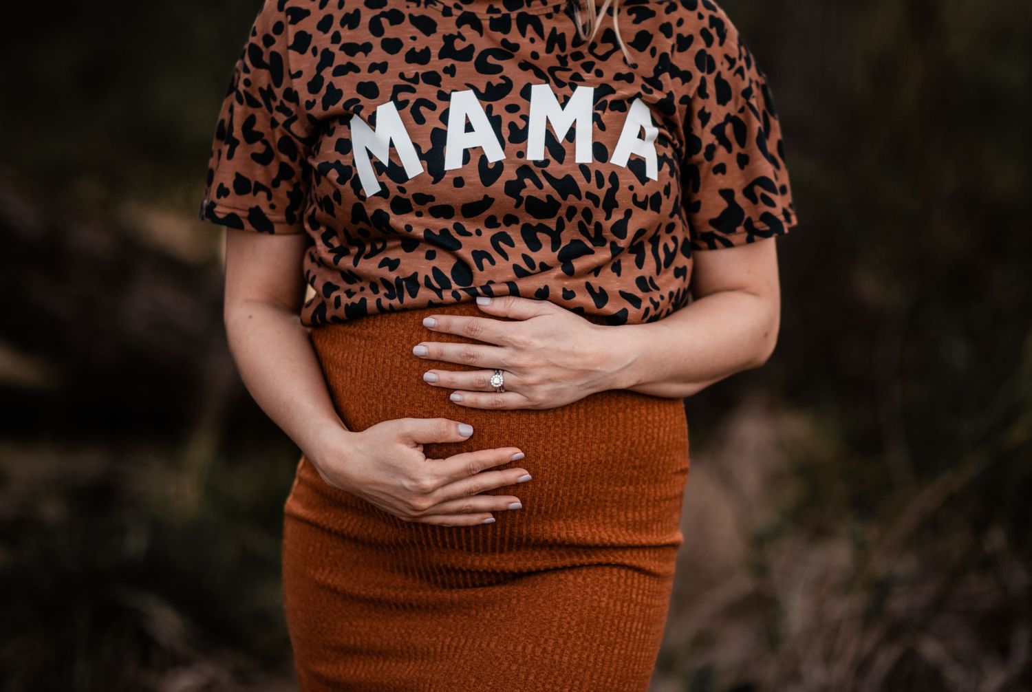 Close up of pregnant belly with leopard print MAMA shirt against dark background.