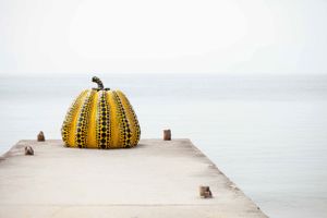Yellow polka dotted pumpkin sculpture by Yayoi Kusama on a concrete pier overlooking water.