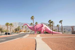 Large pink dinosaur sculpture in desert landscape with palm trees and mountains.