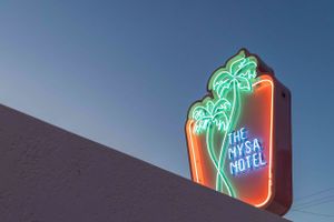 Retro neon The Mysa Motel sign with palm tree design glowing against dark blue sky.