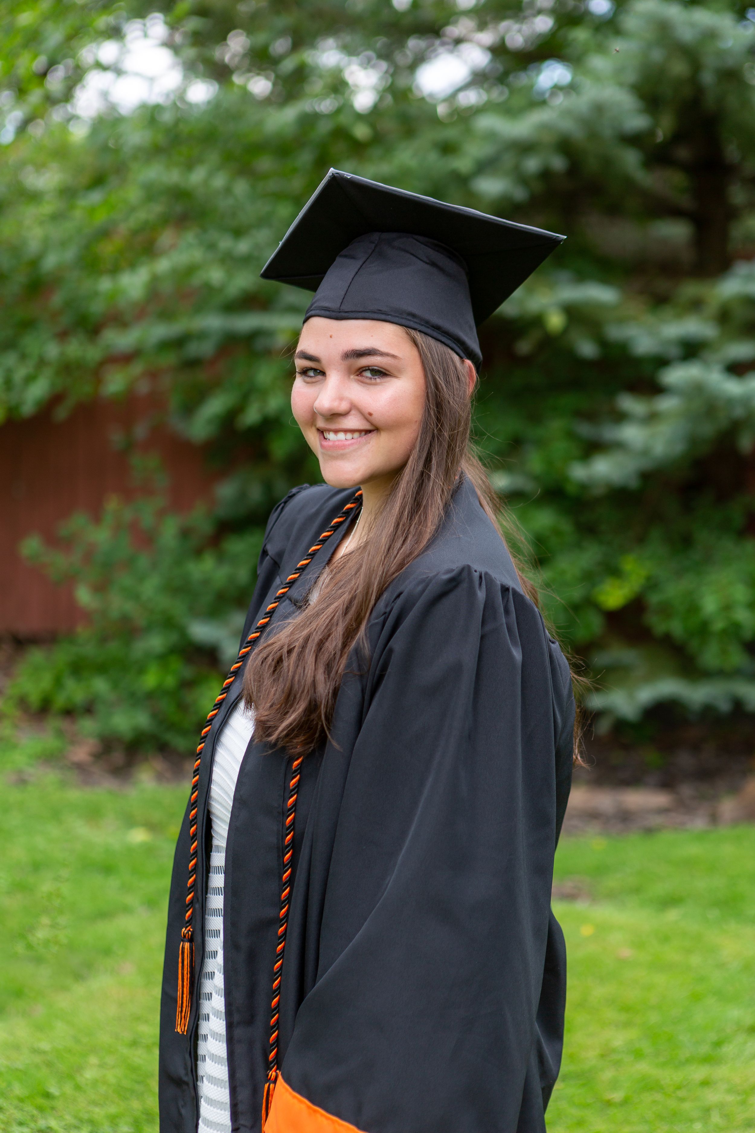 Graduate in black cap and gown with orange honors cord smiles outdoors against green foliage.