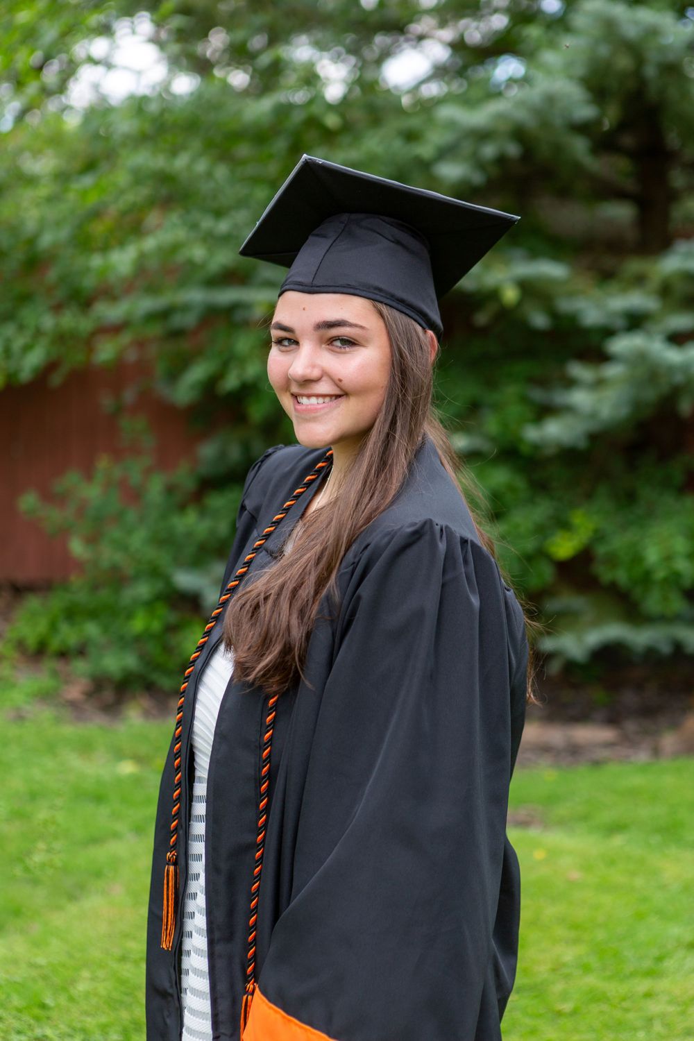Graduate in black cap and gown with orange honors cord smiles outdoors against green foliage.