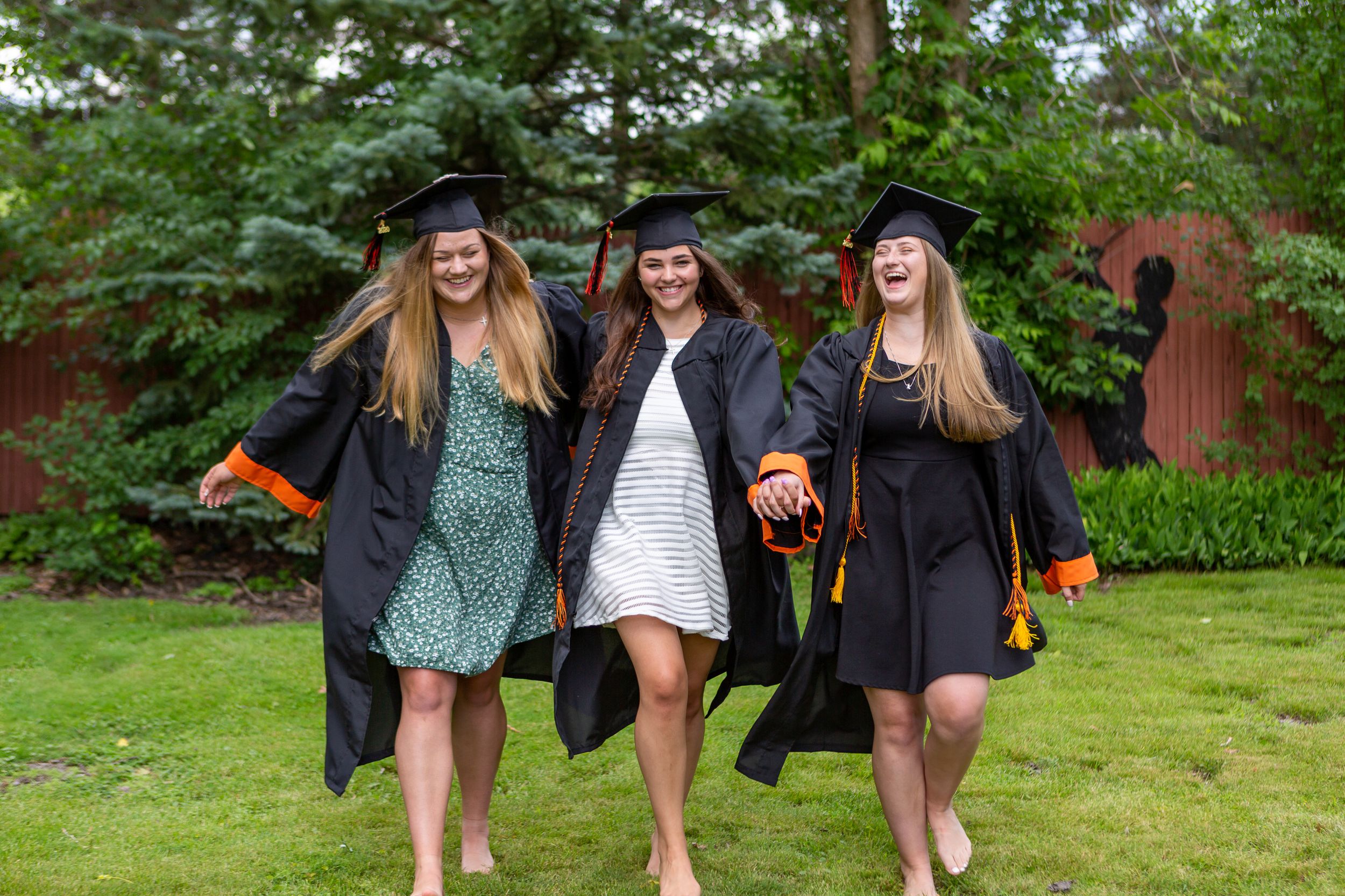 Three graduates in black caps and gowns celebrate together on a lawn.