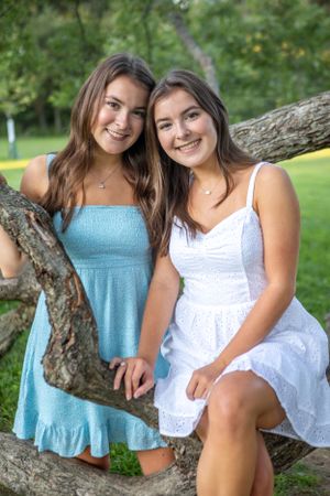 Two friends in summer dresses pose together on a tree branch in a sunny park.