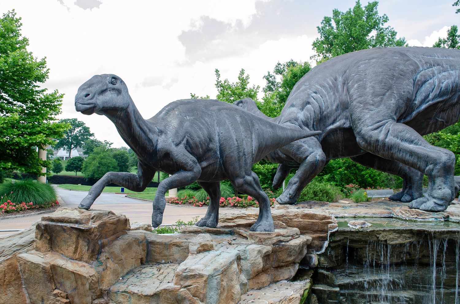 Life-sized dinosaur statues displayed on rocky waterfall feature in outdoor setting.
