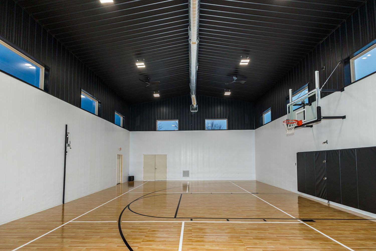 Indoor basketball court with wooden flooring, white walls, and high windows providing natural light.